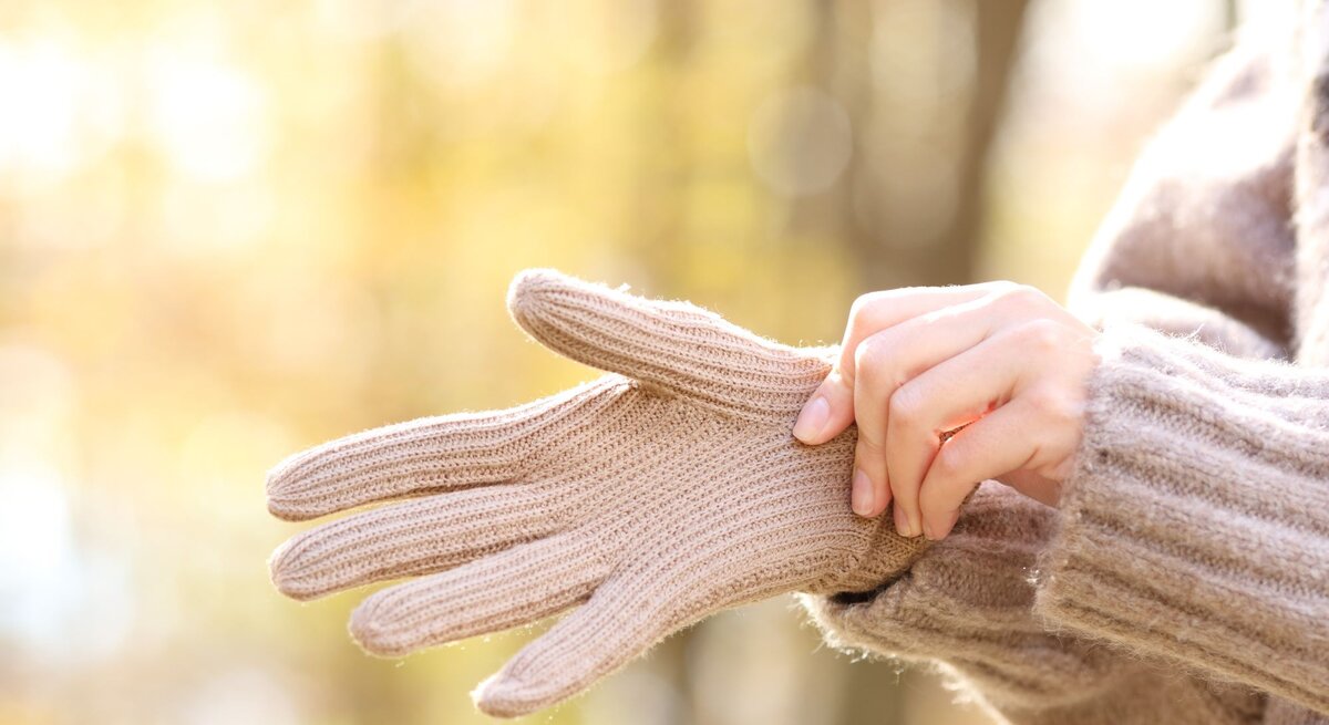 Eine Person zieht sich bei warmem Herbstlicht beige Strickhandschuhe über, im Hintergrund ein Wald in sanften Unschärfen.