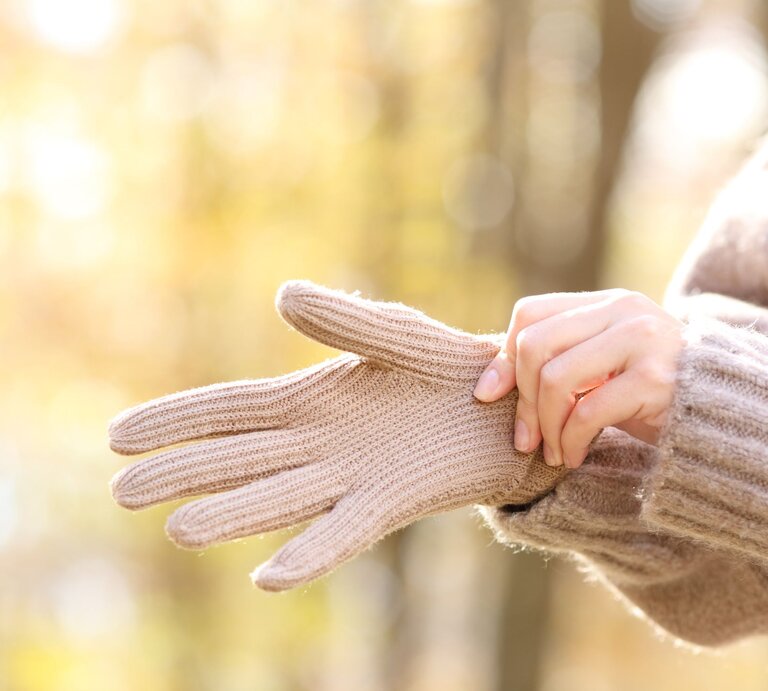 Eine Person zieht sich bei warmem Herbstlicht beige Strickhandschuhe über, im Hintergrund ein Wald in sanften Unschärfen. | © Adobe Stock/Antonioguillem