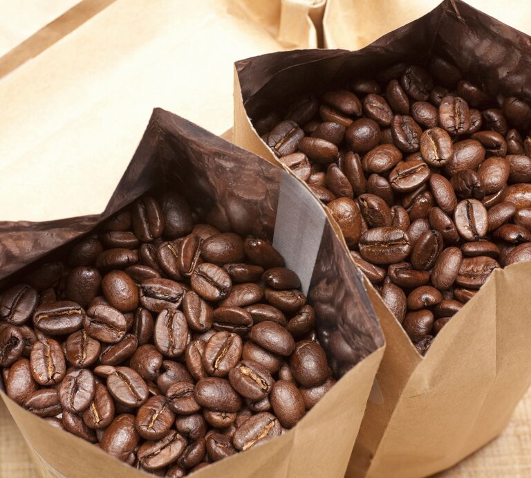 Zwei geöffnete Papiertüten, randvoll mit glänzenden, dunkelbraunen Kaffeebohnen, auf einem Holztisch liegend. | © Adobe Stock/Paylessimages