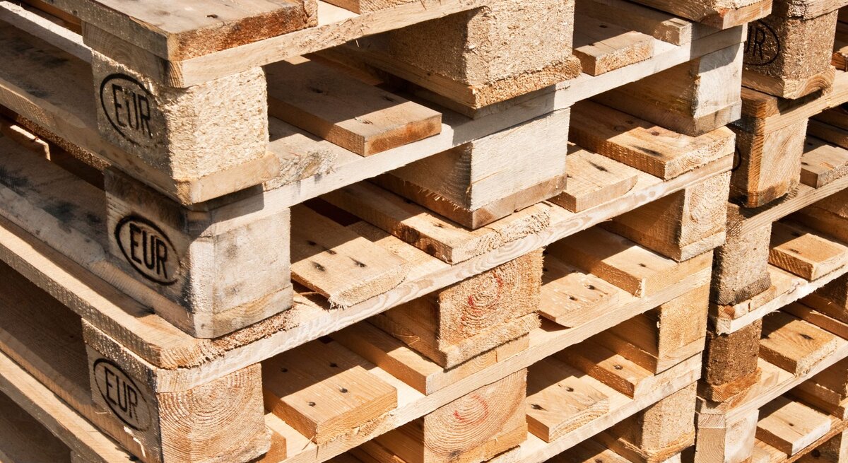 Stapel aus hellem Holz gefertigten Europaletten mit sichtbarem EUR-Logo, teils mit Gebrauchsspuren, in warmem Sonnenlicht aufgenommen.