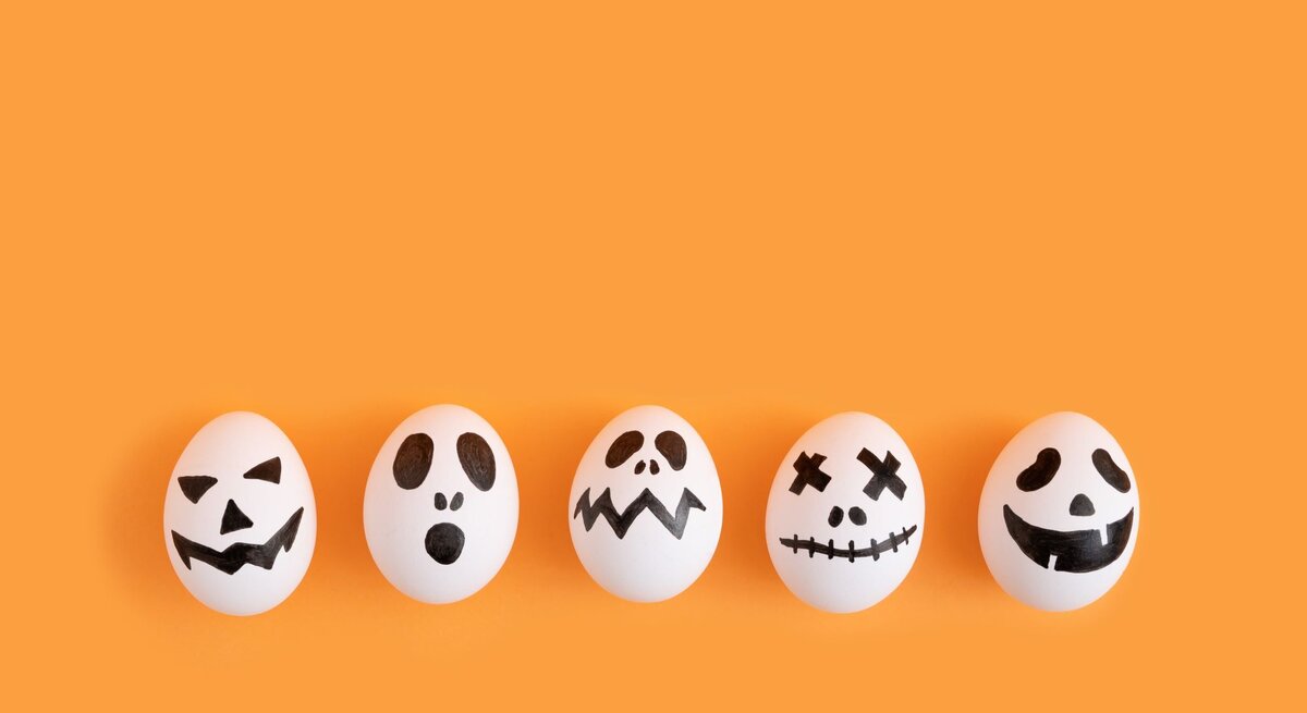 Fünf weiße Eier mit gruseligen schwarzen Halloween-Gesichtern liegen nebeneinander vor einem orangefarbenen Hintergrund.