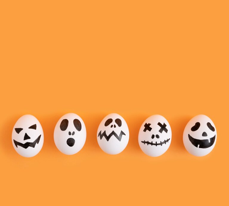 Fünf weiße Eier mit gruseligen schwarzen Halloween-Gesichtern liegen nebeneinander vor einem orangefarbenen Hintergrund. | © AdobeStock/Drpixel
