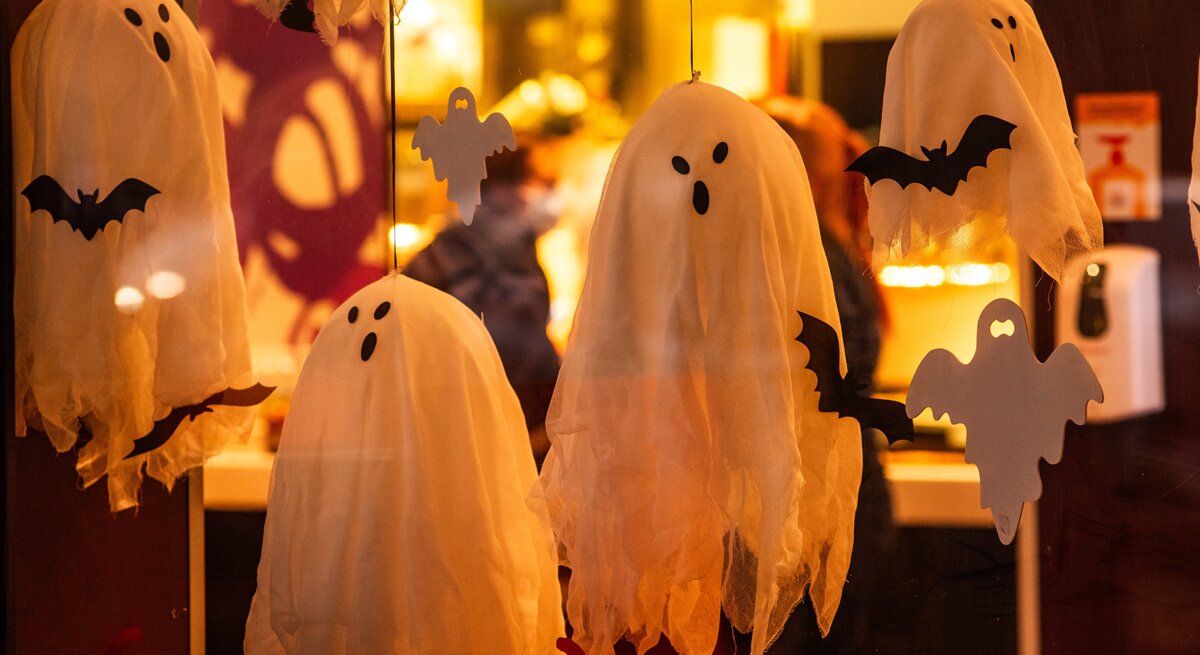 Weiße Geister und schwarze Fledermäuse aus Stoff hängen als Halloween-Dekoration hinter einer erleuchteten Glasscheibe.