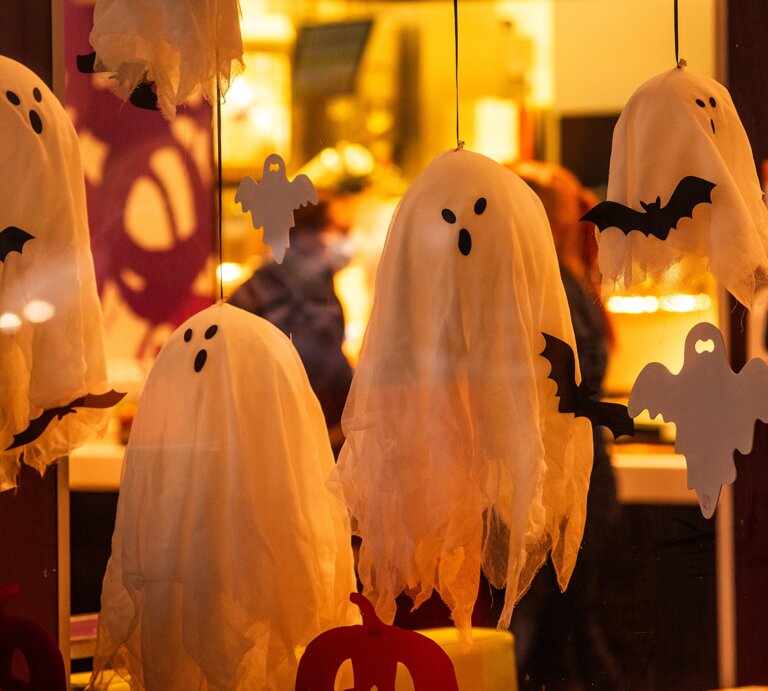 Weiße Geister und schwarze Fledermäuse aus Stoff hängen als Halloween-Dekoration hinter einer erleuchteten Glasscheibe. | © GettyImages/Yuliia Pavaliuk