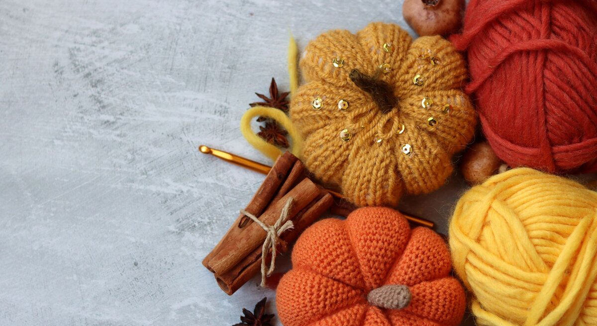 Herbstliche Deko mit orangefarbenen Häkelkürbissen, glänzender Wolle, Zimt, Sternanis und goldener Häkelnadel auf Betonhintergrund.