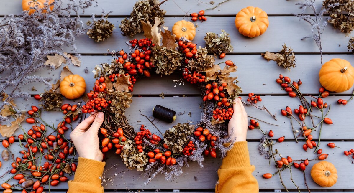 Zwei Hände in senfgelbem Pullover halten einen herbstlichen Kranz aus Hagebutten, Trockenblüten und Eichenblättern auf Holztisch.
