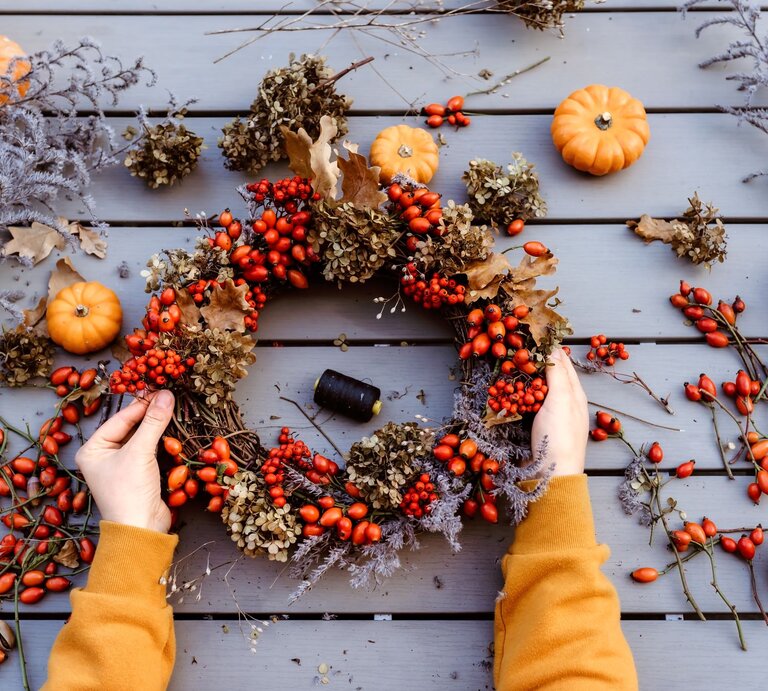 Zwei Hände in senfgelbem Pullover halten einen herbstlichen Kranz aus Hagebutten, Trockenblüten und Eichenblättern auf Holztisch. | © Shutterstock/Tatyana Soares