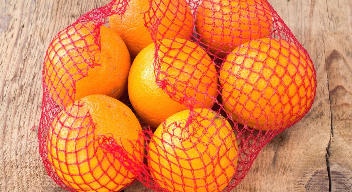 Ein rotes Netz mit sieben frischen Orangen liegt auf rustikalem Holzuntergrund.