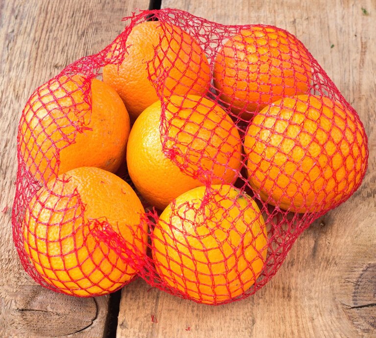 Ein rotes Netz mit sieben frischen Orangen liegt auf rustikalem Holzuntergrund. | © Adobe Stock/gavran333