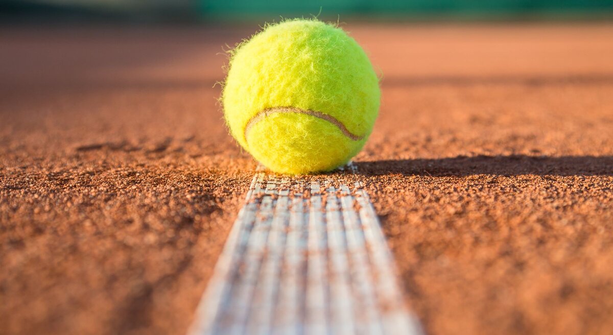 Ein gelber Tennisball liegt knapp über der weißen Linie auf einem sandigen Tennisplatz in der Sonne.
