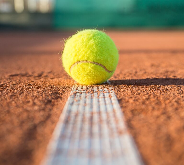 Ein gelber Tennisball liegt knapp über der weißen Linie auf einem sandigen Tennisplatz in der Sonne. | © Adobe Stock/pablobenii