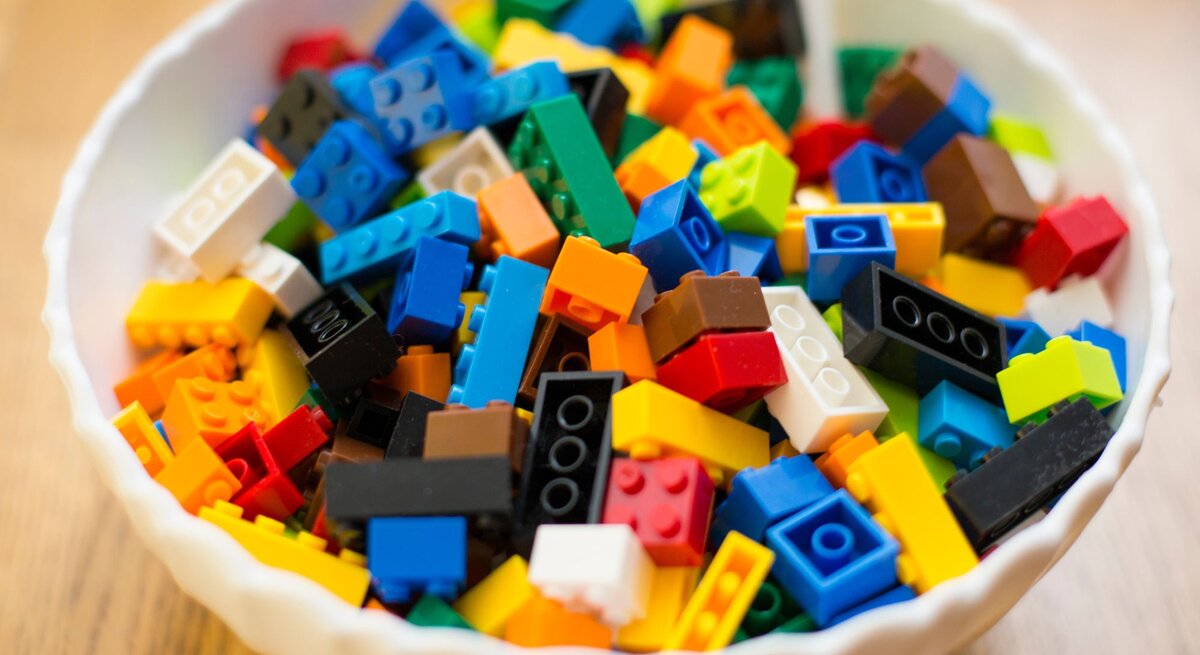Eine weiße Schale voller bunter Legosteine in verschiedenen Formen und Farben steht auf einem Holztisch.