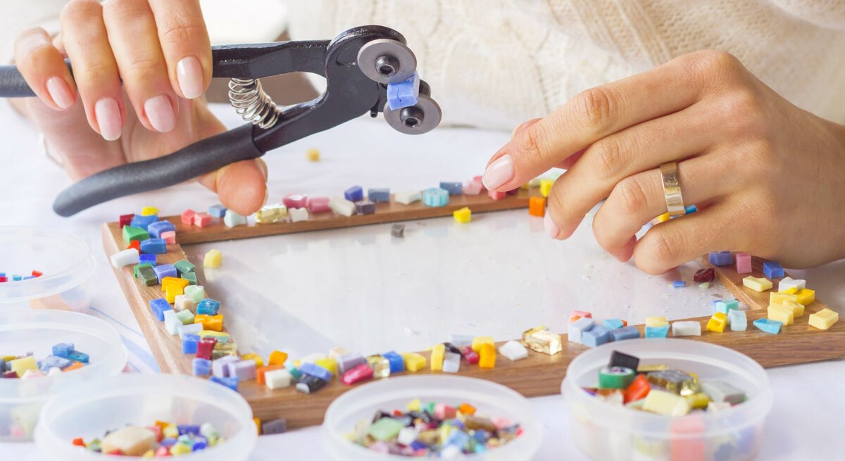 Hände einer Person setzen bunte Mosaiksteine mit einer Zange auf einen Bilderrahmen, umgeben von Schälchen mit Mosaikteilen.