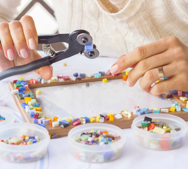 Hände einer Person setzen bunte Mosaiksteine mit einer Zange auf einen Bilderrahmen, umgeben von Schälchen mit Mosaikteilen. | © Adobe Stock/Evgeniya
