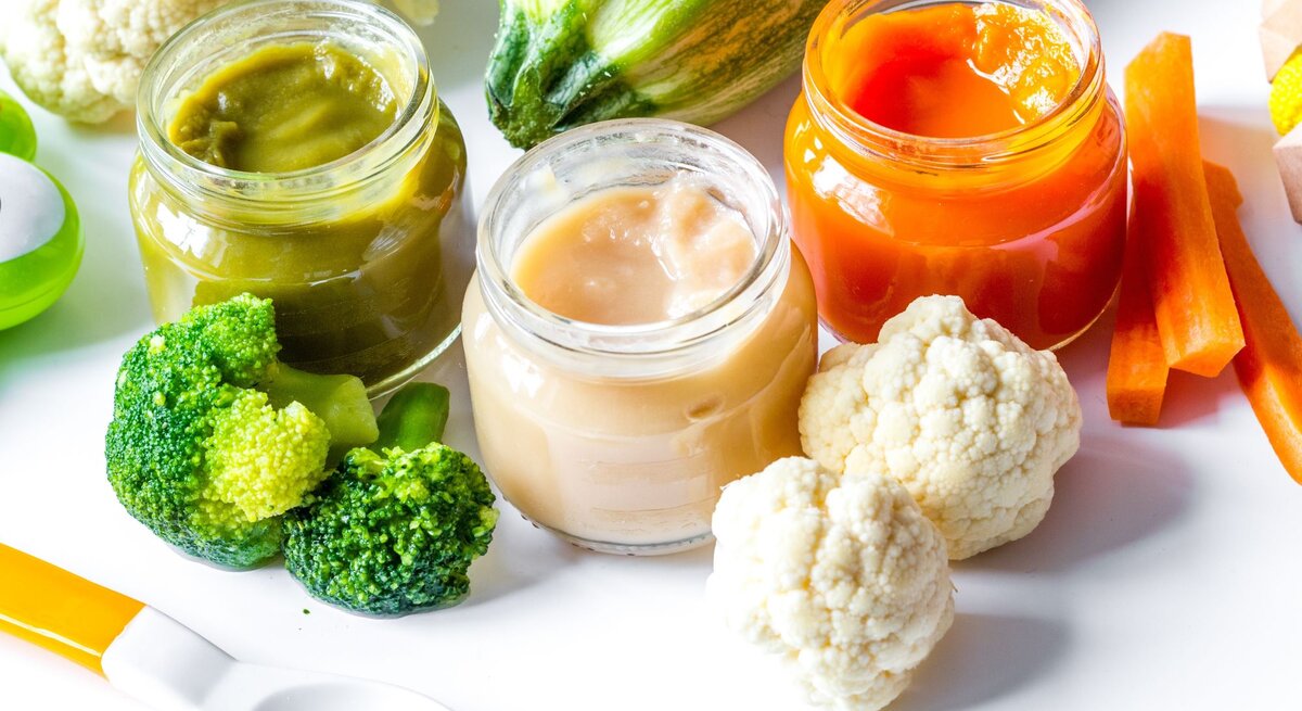 Drei offene Gläser mit Babybrei in Grün, Beige und Orange stehen auf einem weißen Tisch, umgeben von frischem Brokkoli, Blumenkohl, Karotten und Zucchini.