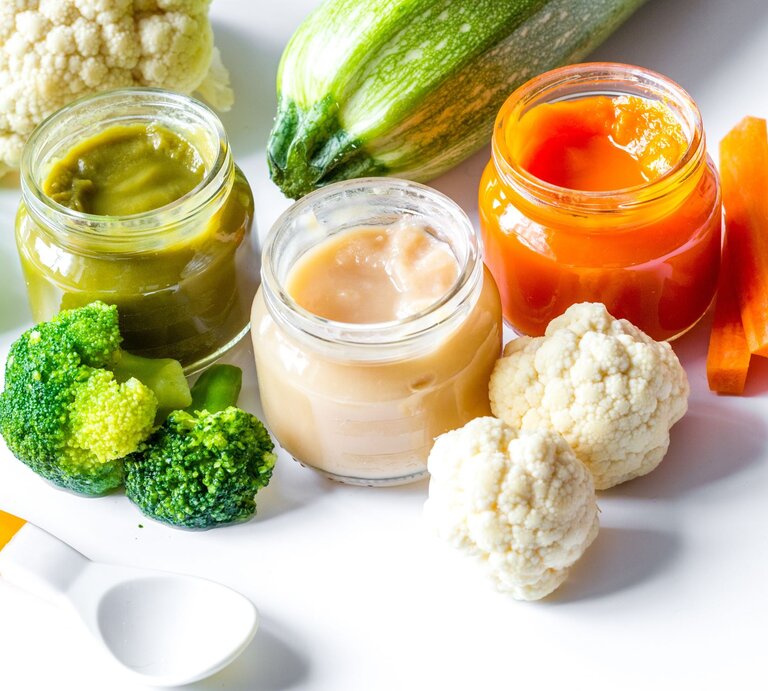 Drei offene Gläser mit Babybrei in Grün, Beige und Orange stehen auf einem weißen Tisch, umgeben von frischem Brokkoli, Blumenkohl, Karotten und Zucchini. | © Adobe Stock/279photo
