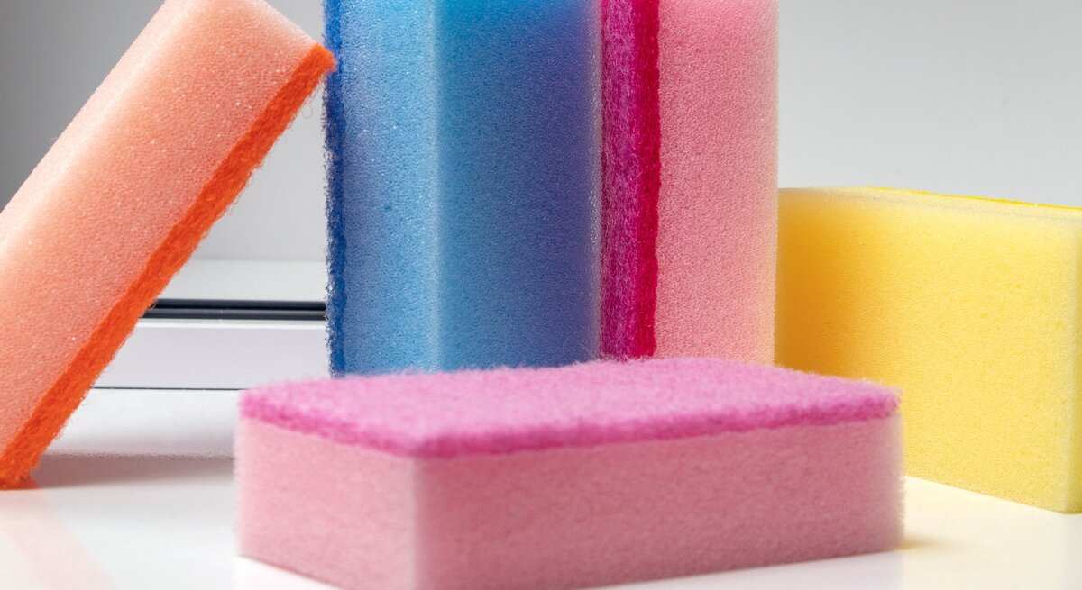 Bunte Reinigungsschwämme in Gelb, Blau, Rosa und Orange stehen aufrecht und liegen auf einem weißen Tisch.