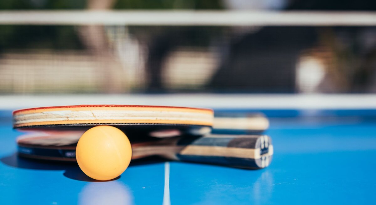 Zwei Tischtennisschläger und ein Ball liegen auf einem blauen Tisch vor unscharfem Hintergrund im Freien.