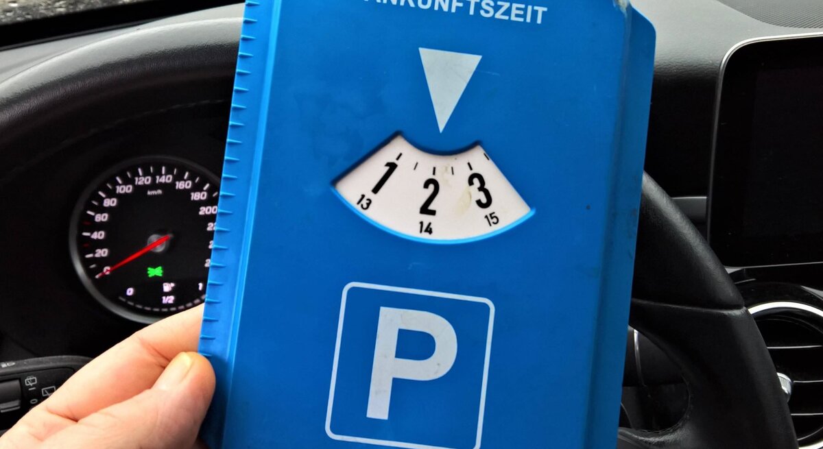 Eine Hand hält eine blaue Parkscheibe mit der Ankunftszeit auf 14 Uhr vor einem Autolenkrad und einem Tacho.