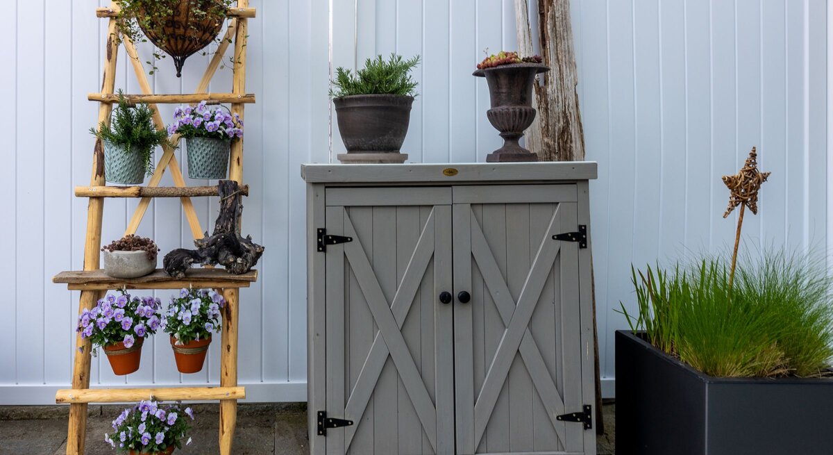 Blühende violette Blumen, Tontöpfe und Kräuter arrangiert auf Leiterregal neben rustikalem Gartenschrank aus Holz.