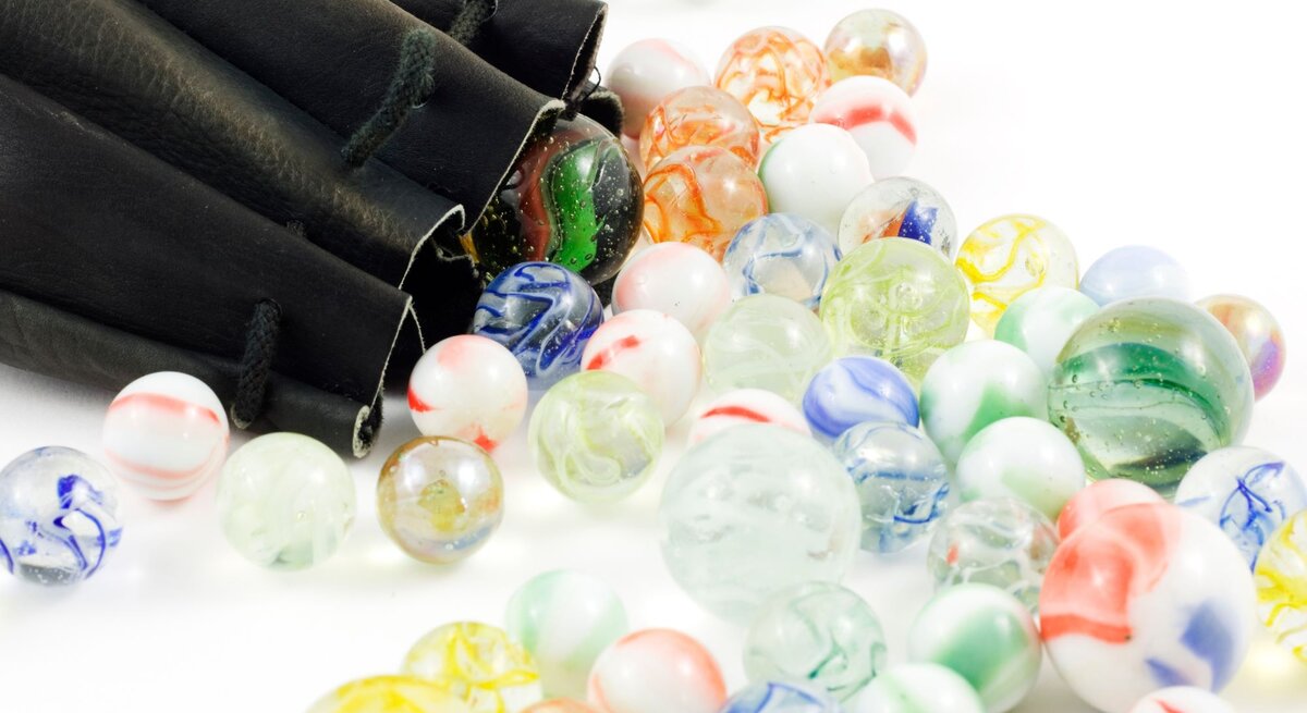 Bunte Glasmurmeln mit marmorierten Mustern rollen aus einem schwarzen Ledersäckchen auf eine weiße Fläche.