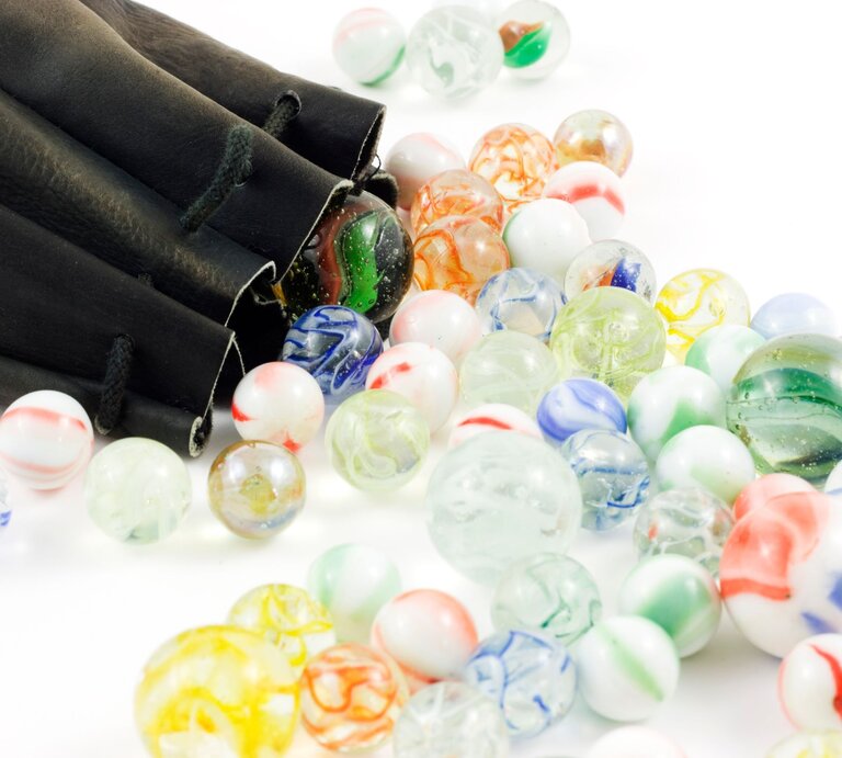 Bunte Glasmurmeln mit marmorierten Mustern rollen aus einem schwarzen Ledersäckchen auf eine weiße Fläche. | © Adobe Stock/soulrebel83