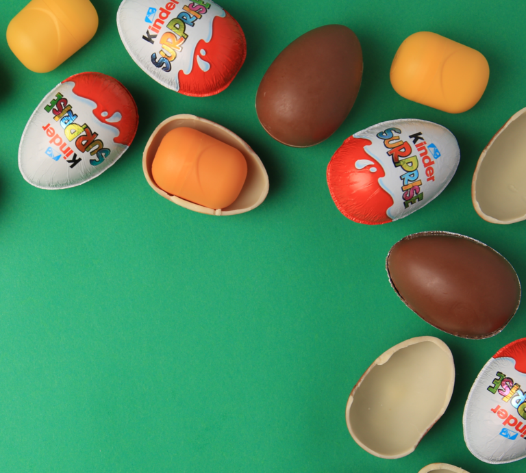 Mehrere bunte Überraschungseier, teils geöffnet mit gelben Kapseln, liegen verstreut auf grünem Hintergrund. | © canva