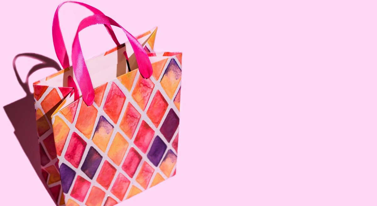 Bunte Geschenktüte mit pinken Griffen und rautenförmigem Muster in Rot-, Orange- und Lilatönen auf rosa Hintergrund.