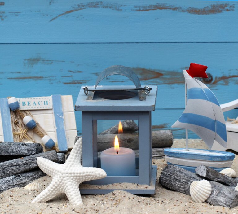 Maritime Deko mit blauer Laterne, kleiner Segelbootfigur, Muscheln, Seestern und dekorativen Holzstücken vor blauer Holzwand. | © Adobe Stock/Racamani