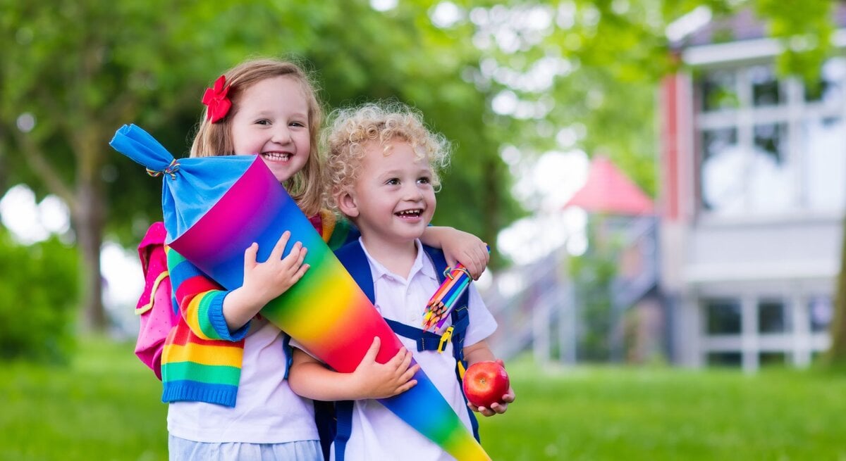 Zwei fröhliche Kinder, eines mit Locken und eines mit Haarspange, halten eine bunte Schultüte im Grünen vor einem Schulgebäude.