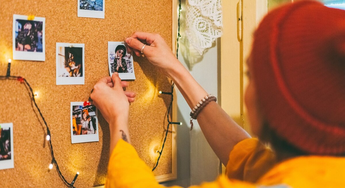 Junge Frau mit roter Mütze und gelbem Pullover pinnt Polaroid-Foto an eine Pinnwand aus Kork mit Lichterkette.