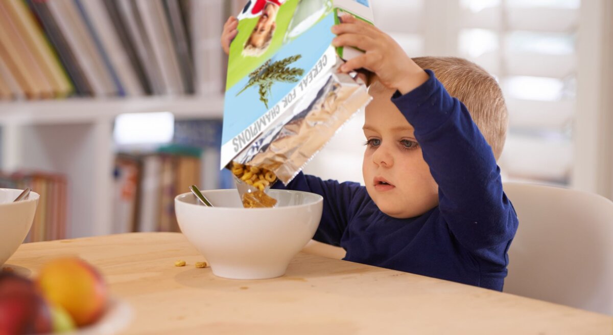 Ein kleiner Junge in blauem Pullover füllt konzentriert Müsli aus einer bunten Packung in eine weiße Schüssel am Holztisch.