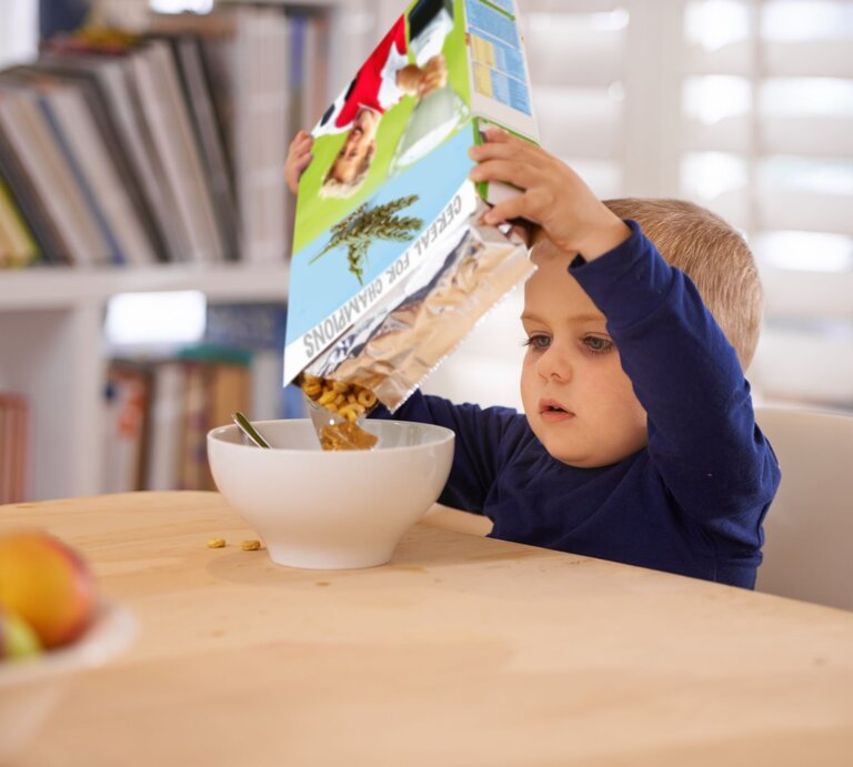Ein kleiner Junge in blauem Pullover füllt konzentriert Müsli aus einer bunten Packung in eine weiße Schüssel am Holztisch. | © Getty Images/PeopleImages