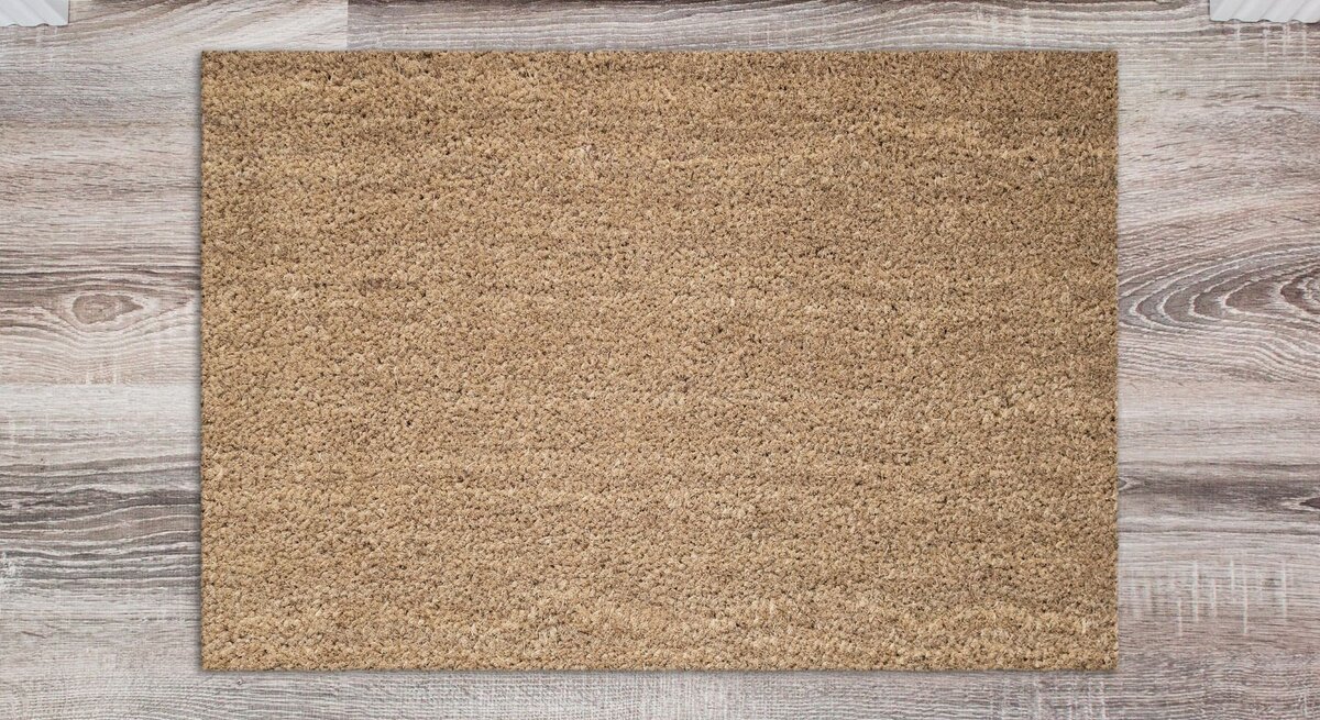 Rechteckige beige Fußmatte auf hellem Holzfußboden vor weißer Eingangstür mit Fenstern.