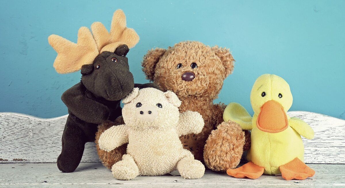 Vier bunte Stofftiere, darunter ein Elch, ein Teddy, ein Schweinchen und eine Ente, sitzen vor einer blauen Wand auf hellem Holzboden.