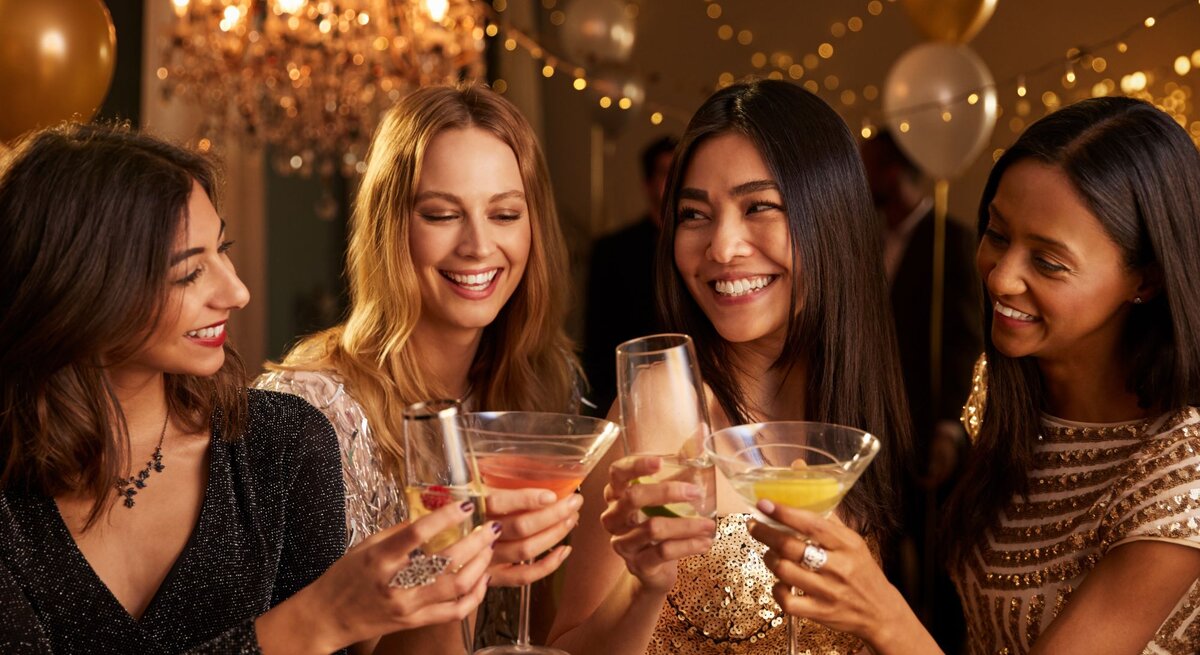 Vier junge Frauen in glitzernden Abendkleidern stoßen lachend mit Cocktails in einem festlich beleuchteten Raum an.