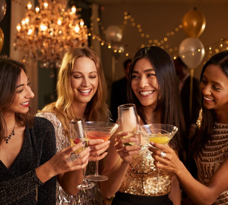 Vier junge Frauen in glitzernden Abendkleidern stoßen lachend mit Cocktails in einem festlich beleuchteten Raum an. | © Adobe Stock/Monkey Business
