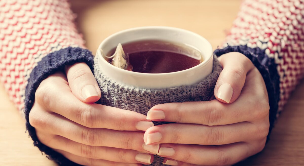 Zwei Hände mit zarten, lackierten Fingernägeln umschließen eine Teetasse mit Teebeutel darin mit gestricktem Überzug auf einem Holztisch.