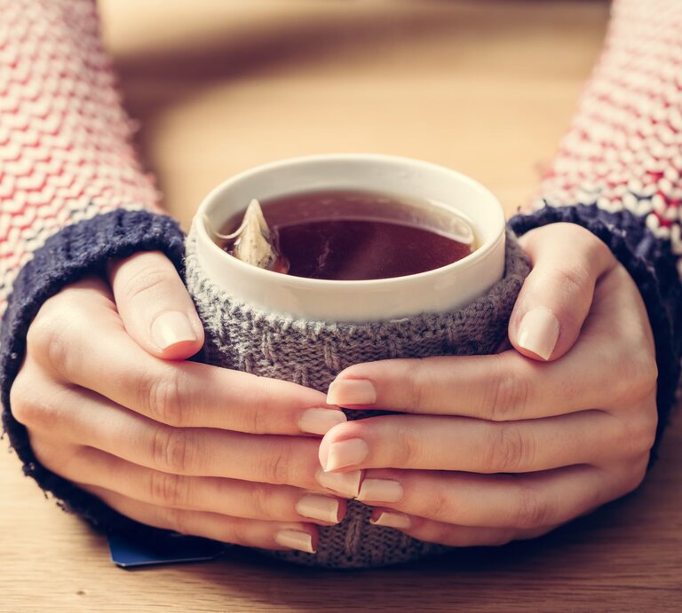 Zwei Hände mit zarten, lackierten Fingernägeln umschließen eine Teetasse mit Teebeutel darin mit gestricktem Überzug auf einem Holztisch. | © Adobe Stock/Photocreo Bednarek