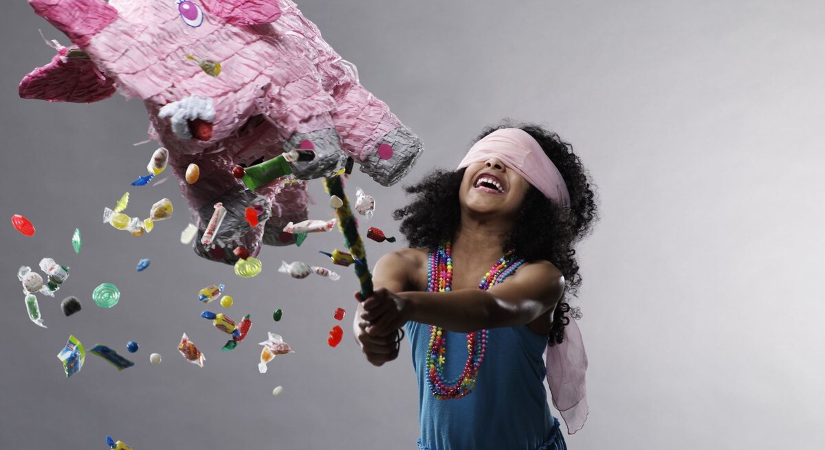 Mädchen mit lockigem Haar und bunter Halskette zerschlägt fröhlich eine rosa Elefanten-Piñata, aus der Bonbons herausfallen.