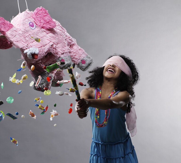 Mädchen mit lockigem Haar und bunter Halskette zerschlägt fröhlich eine rosa Elefanten-Piñata, aus der Bonbons herausfallen. | © Getty Images/Ryan McVay