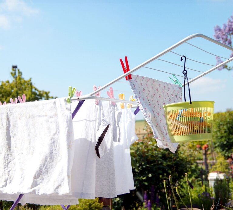 Weiße Wäsche und ein gemustertes Tuch hängen auf einem Wäscheständer im sonnigen Garten mit blühenden Bäumen. | © Adobe Stock/ImageSine