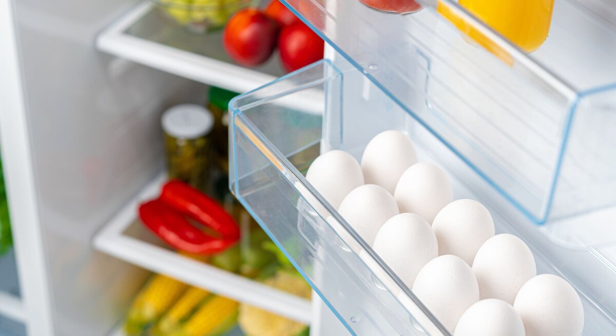 Ein geöffneter Kühlschrank zeigt Eier, Säfte und frisches Gemüse wie Paprika und Mais in den Regalen.