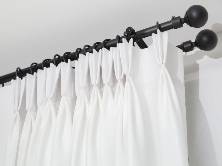 Ein weißer Vorhang mit Falten hängt mithilfe von Gardinenringen an einer schwarzen Gardinenstange mit Kugelenden vor einer weißen Wand. | © Adobe Stock/bennnn