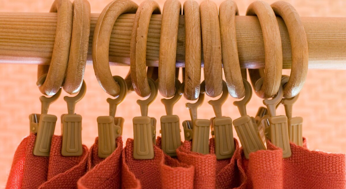 Nahaufnahme einer Holzgardinenstange mit mehreren Gardinenringen aus Holz und beigefarbenen Clips, an denen ein roter Stoffvorhang befestigt ist.