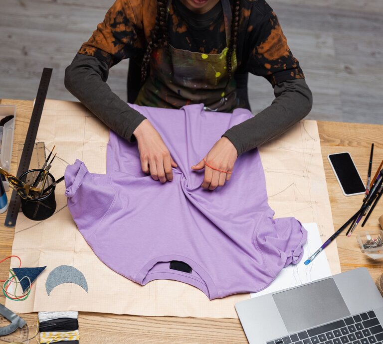 Kreative Arbeit an einem lila T-Shirt, umgeben von Pinsel, Lineal, Laptop und Bastelmaterialien auf einem Holztisch. | © AdobeStock/LIGHTFIELD STUDIOS
