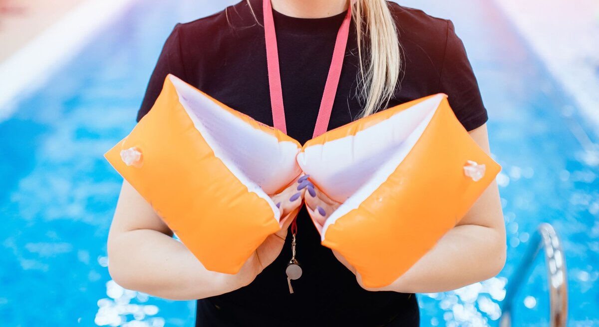Eine blonde Frau in schwarzem T-Shirt hält orange Schwimmflügel vor sich, im Hintergrund ein Schwimmbecken