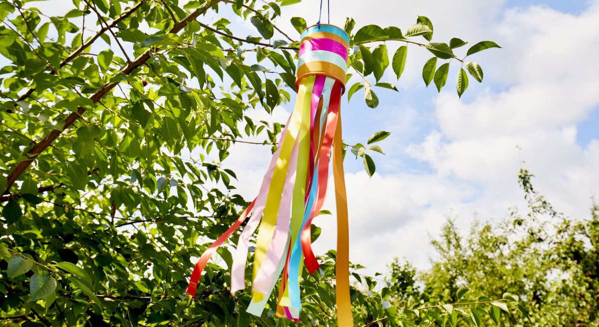 Ein Windspiel aus bunten Bändern weht im Sommerwind an einem grünen Ast unter einem klaren, bewölkten Himmel.
