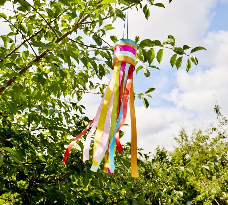Ein Windspiel aus bunten Bändern weht im Sommerwind an einem grünen Ast unter einem klaren, bewölkten Himmel. | © Getty Images/	the_burtons