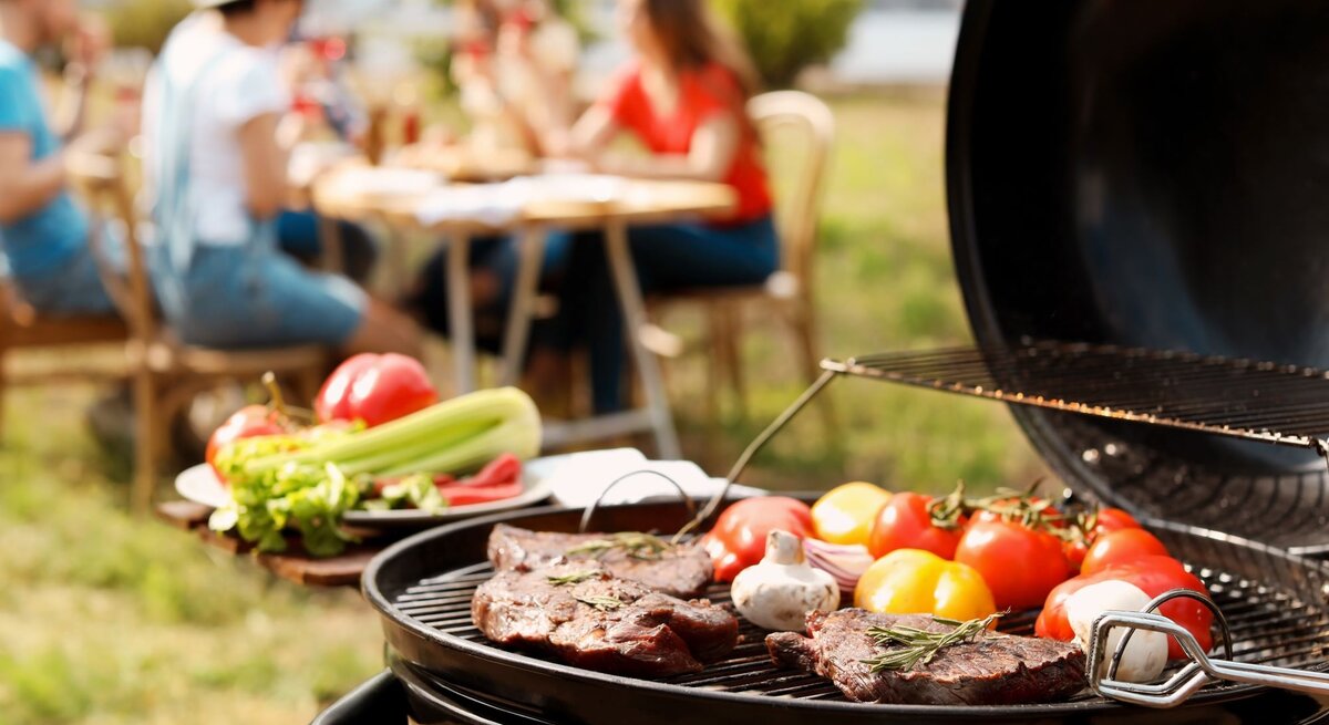 Bunte Gemüse und Steaks werden auf einem Grill zubereitet, während eine Gruppe von Freunden im Hintergrund lacht und isst.